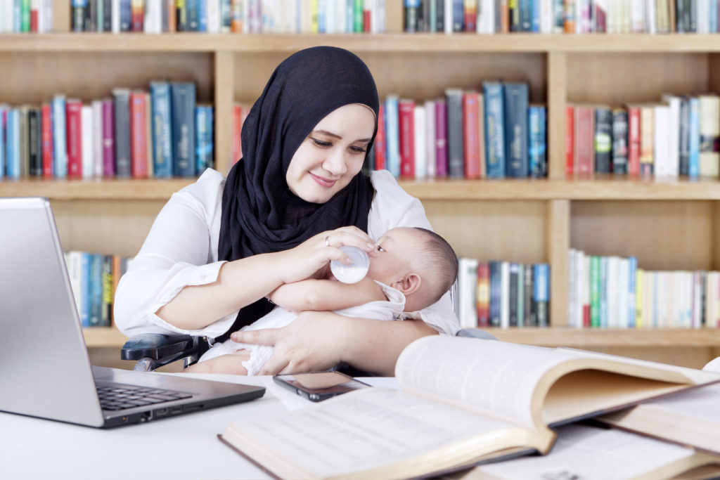 Multitasking young woman in hijab offering her baby milk from bottle while working in the library